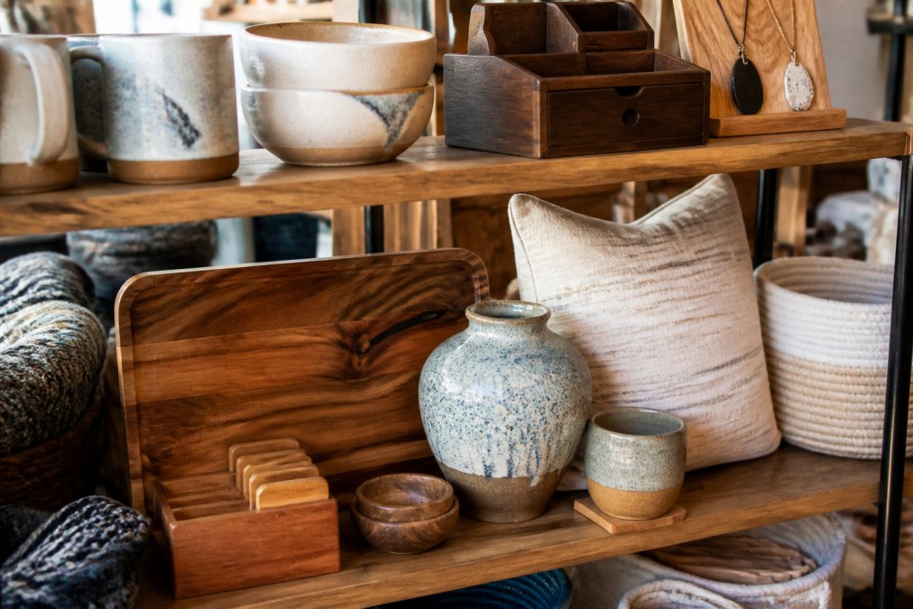 Close-up of artisan-made home accessories and decorative items displayed on a wooden shelf with natural lighting, showing handcrafted pottery, wooden organizers, and personalized items