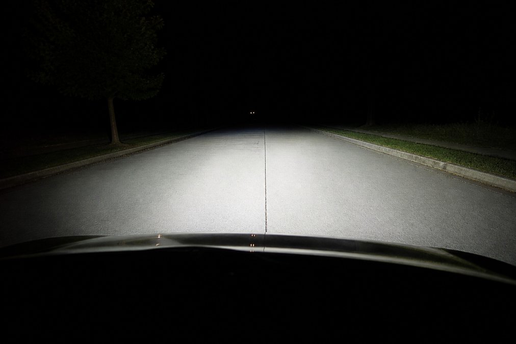 Close-up view of a vehicle's LED headlight beam pattern on a dark road at night, showing a sharp horizontal cutoff line and even illumination coverage across the road surface