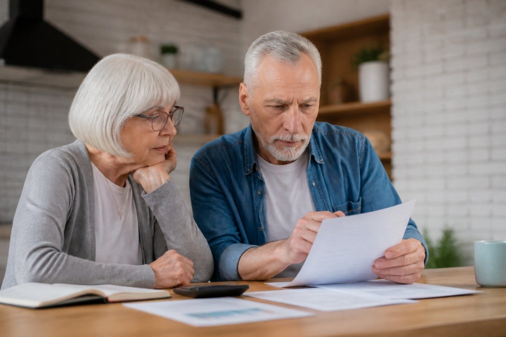 Retired couple reviewing annuity fees and prospectus documents at a kitchen table to understand growth impact.