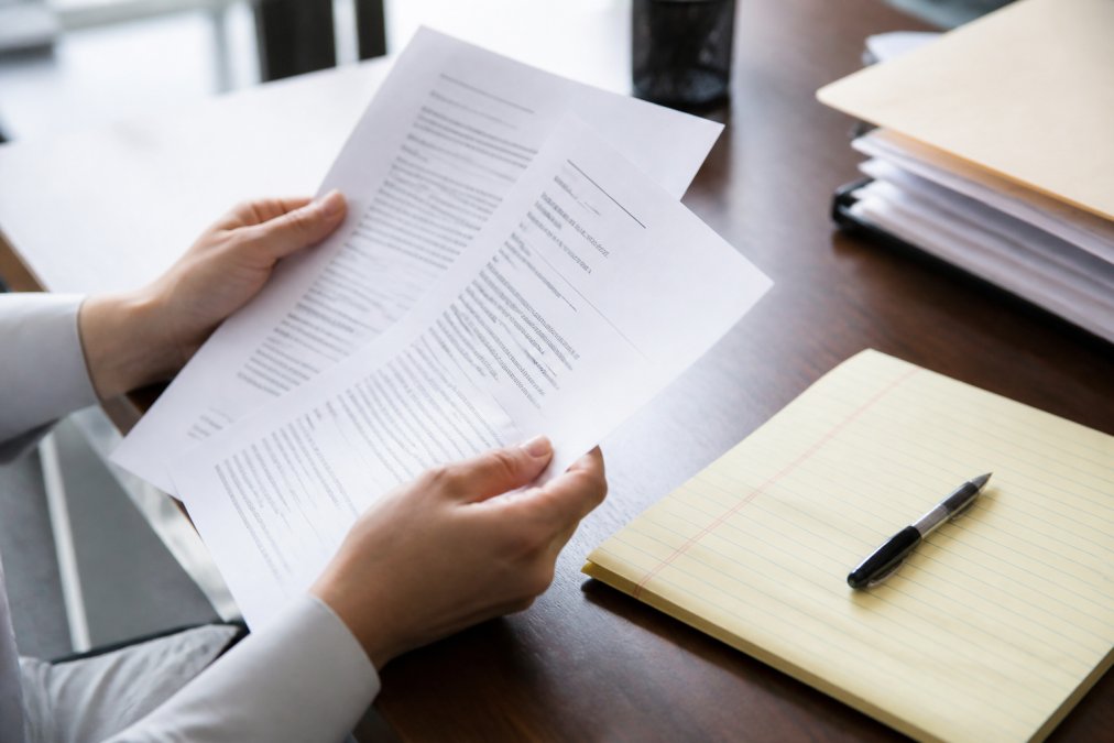 Close-up of hands holding official court documents including a summons and complaint on a desk, with a notepad and pen nearby for taking notes, natural office lighting