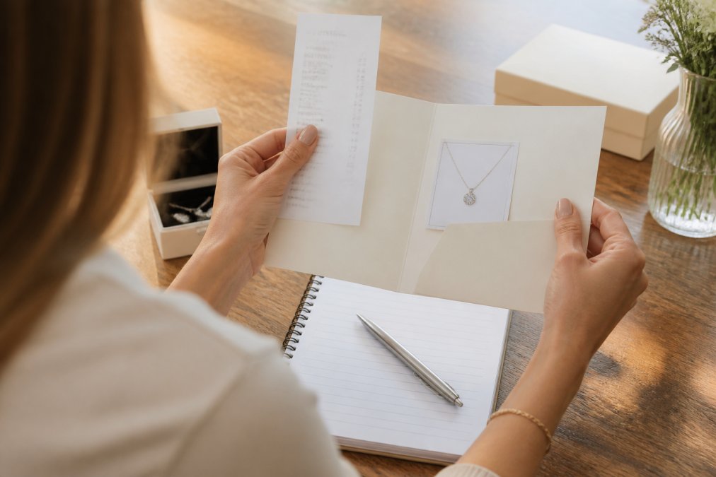 Woman's hands holding jewellery documentation and a receipt on a wooden desk, with a laptop and notepad visible, reviewing purchase records in natural afternoon light