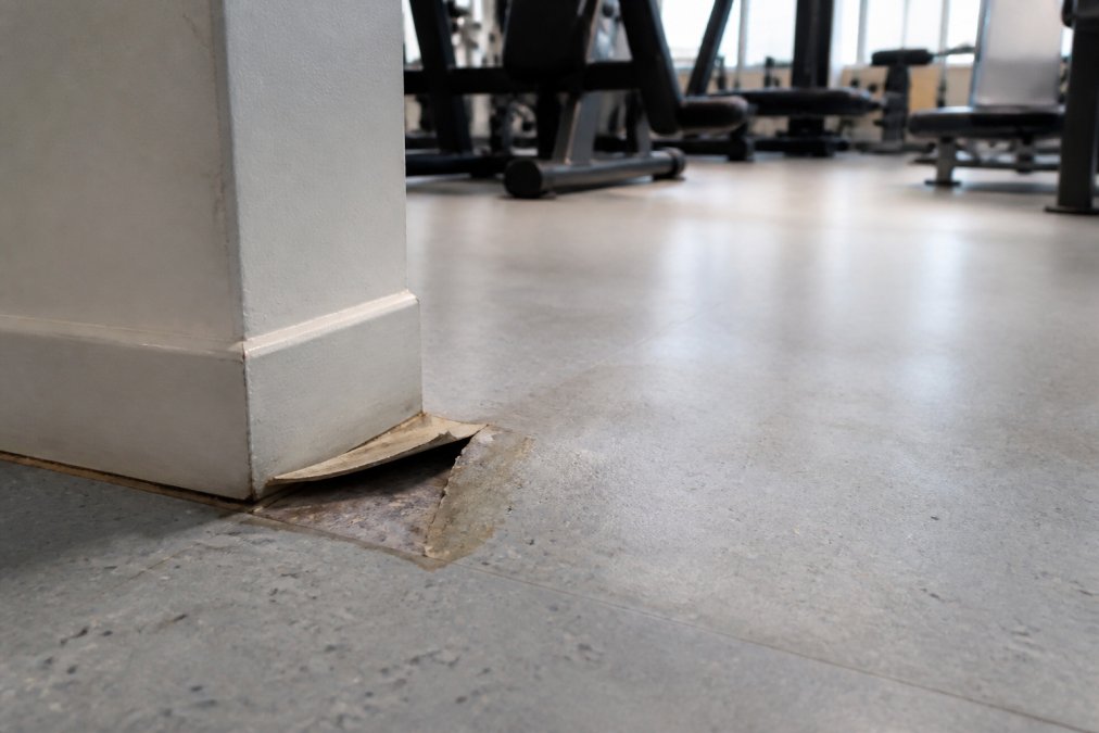 Close-up of a damaged fitness center floor corner with visible discoloration and slight lifting of the vinyl edge, with gym equipment in the blurred background