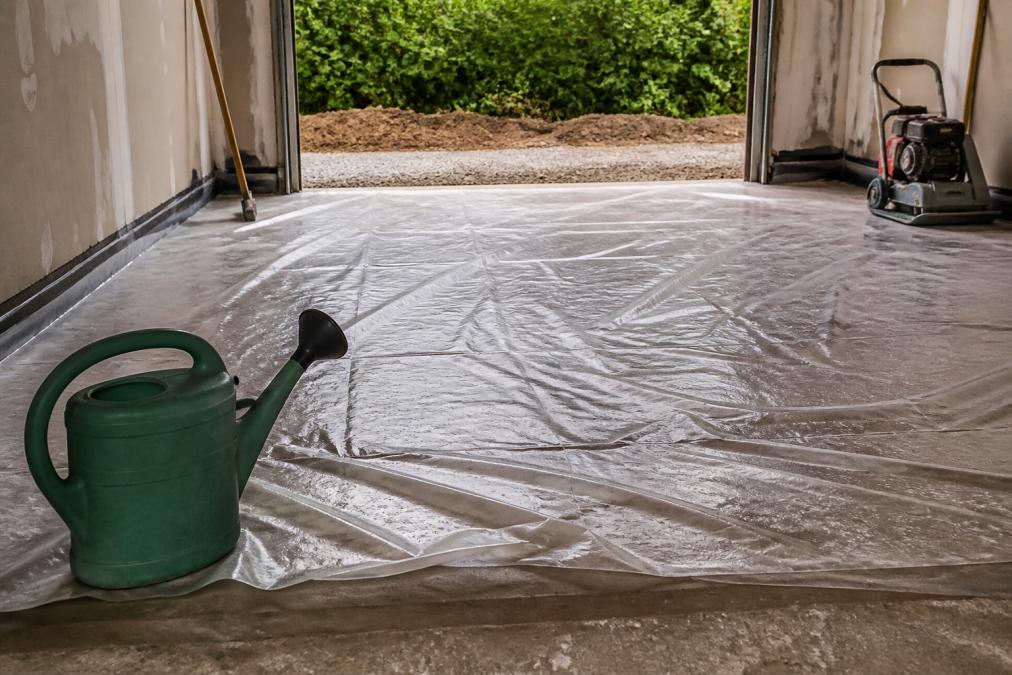 Undgå revner i garagegulv: 7 trin der virker i 2026 2 Close-up of a freshly poured concrete garage floor being covered with plastic sheeting to retain moisture during curing, with a watering can nearby and tools in the background