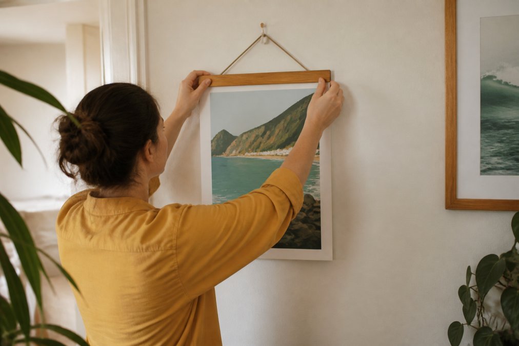 A close-up of hands using a wooden clip hanger to attach an unframed travel poster to a painted wall, with the print visible and no damage to the surface