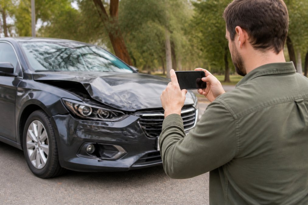 Driver photographing vehicle damage at accident scene with smartphone, capturing license plate and damage clearly visible in daylight, standing beside damaged vehicle