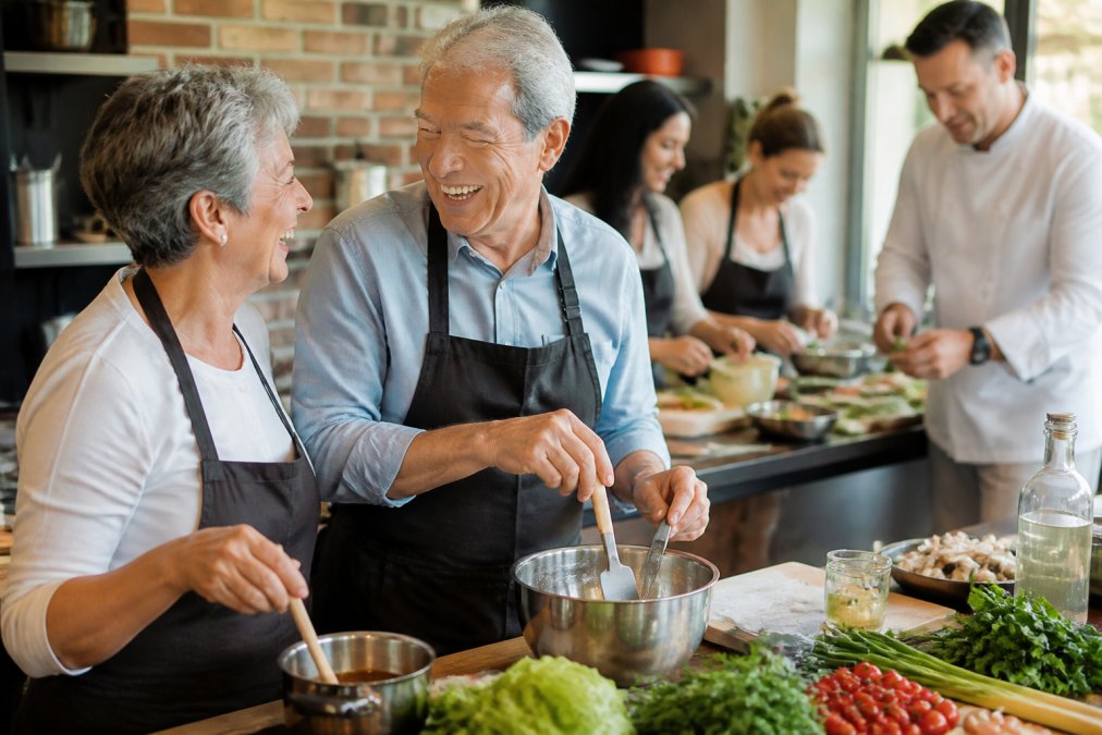 A couple laughing together in a bright cooking class setting, with an instructor demonstrating a dish in the background and other participants visible at their cooking station
