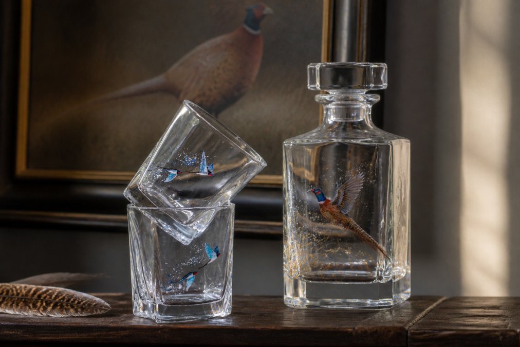 Close-up of hand-painted whisky tumblers and an 800ml decanter featuring richly detailed pheasant artwork, arranged on a dark wooden surface with warm, low natural light filtering from the left