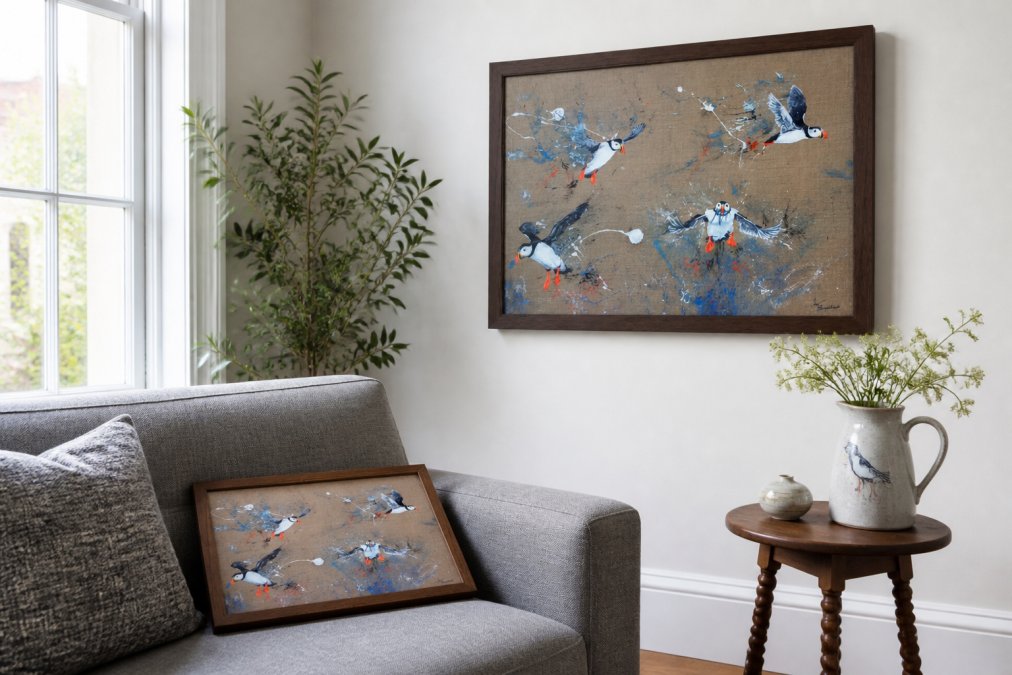 A bright, contemporary living room with a single large framed swallow print on a white wall, a cushioned puffin lap tray resting on a grey linen sofa, and a wooden side table with a hand-painted water jug, natural afternoon light from a large window