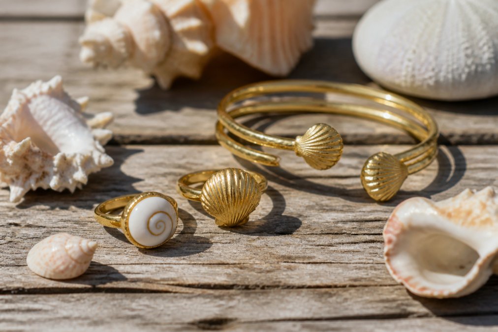 Close-up of coastal jewelry pieces displayed on a weathered wooden surface with seashells and ocean-inspired elements nearby, natural sunlight highlighting the shell textures and gold finishes