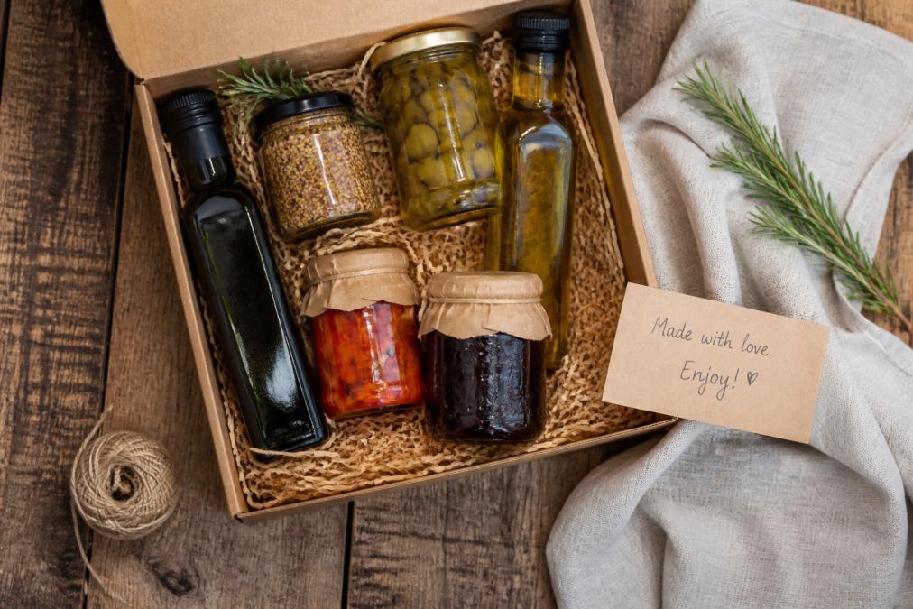 Overhead flat lay of an open artisan food gift box on a rustic wooden table, showing small-batch olive oil bottles, jars of preserves, and a handwritten gift note, with a linen napkin and sprig of rosemary nearby