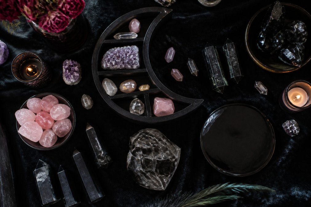 Overhead flat-lay composition of dark aesthetic crystals arranged on a black velvet surface with a crescent moon shelf, rose quartz pieces, black tourmaline towers, and obsidian scrying plates creating a mystical vignette with warm candlelight