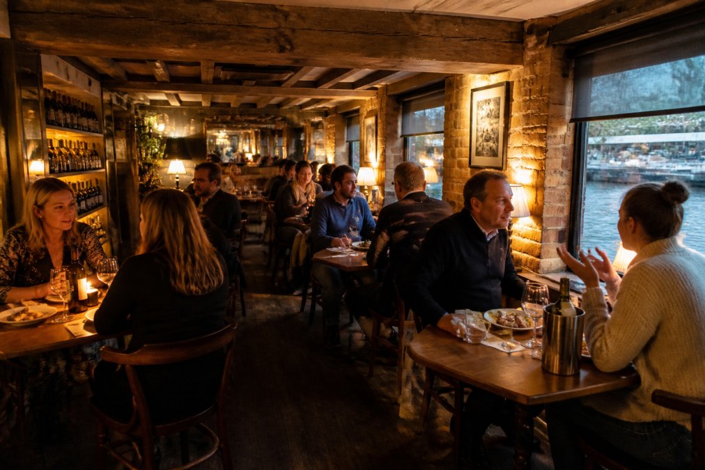 Cosy riverside wine bar interior with warm amber lighting, exposed wooden beams, and guests seated at intimate tables with wine glasses and plates of home-cooked food, river views visible through windows