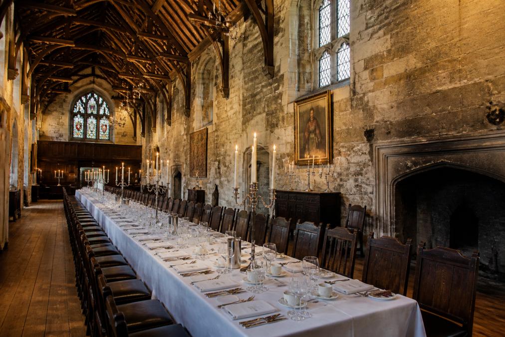 Grand medieval hall with soaring timber-framed ceiling, exposed ancient stone walls, and a long dining table elegantly set with white linens, place settings, and candlelight casting warm shadows on historic architecture