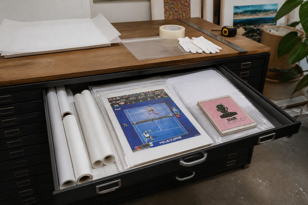 Neatly organized flat file drawer partially open revealing rolled and flat prints in acid-free sleeves, studio environment with soft overhead lighting