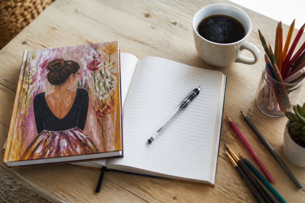 Overhead flat lay of an open hardcover notebook with colorful illustrated cover, pen resting on lined pages, with a steaming cup of coffee and creative supplies including colored pencils scattered nearby on a light wooden desk
