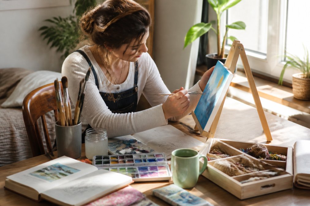 A person sitting at a sunlit table, painting a small canvas while surrounded by colorful art supplies, sketchbooks, and a half-finished DIY craft kit
