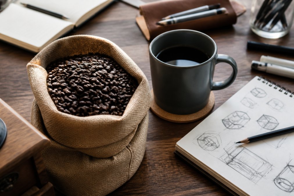 Close-up of premium coffee beans in a stylish container next to a designer's workspace, with a warm mug and design sketches visible