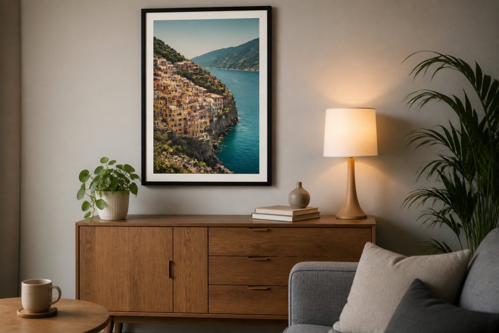 A framed travel poster displayed on a living room wall above a wooden sideboard, with soft ambient lighting, a small potted plant, and minimal Scandinavian-style decor arranged around it