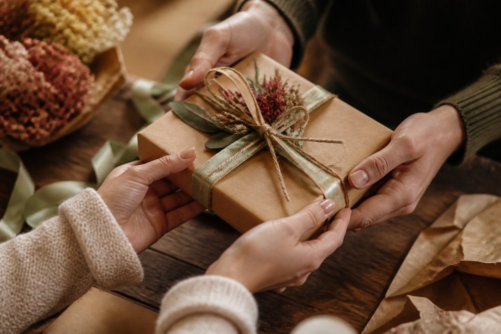 Close-up of hands exchanging a beautifully wrapped artisanal gift box with warm lighting, tissue paper, and natural wooden surface background