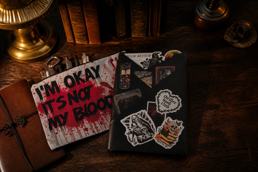 A wrapped hardcover mystery book with leather journal, quality pens, bookmarks, and sticky notes artfully arranged on a dark wooden table with moody lamplight casting shadows, suggesting anticipation and discovery