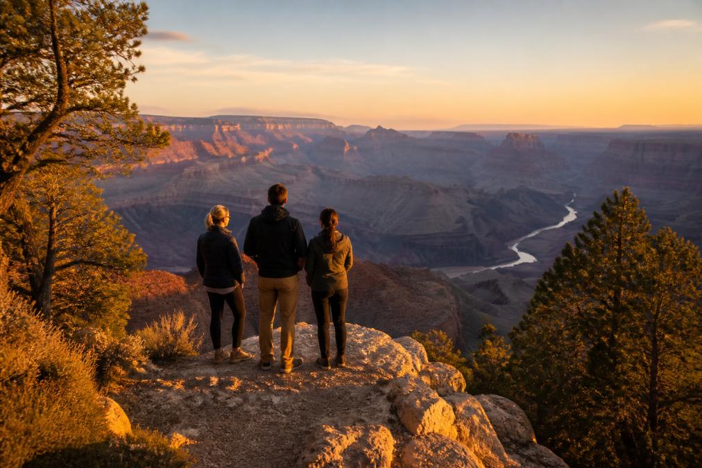 A small group of travelers standing at a quiet Grand Canyon South Rim overlook at golden hour, with layered rock formations and the Colorado River visible far below, pine trees framing the edge of the canyon