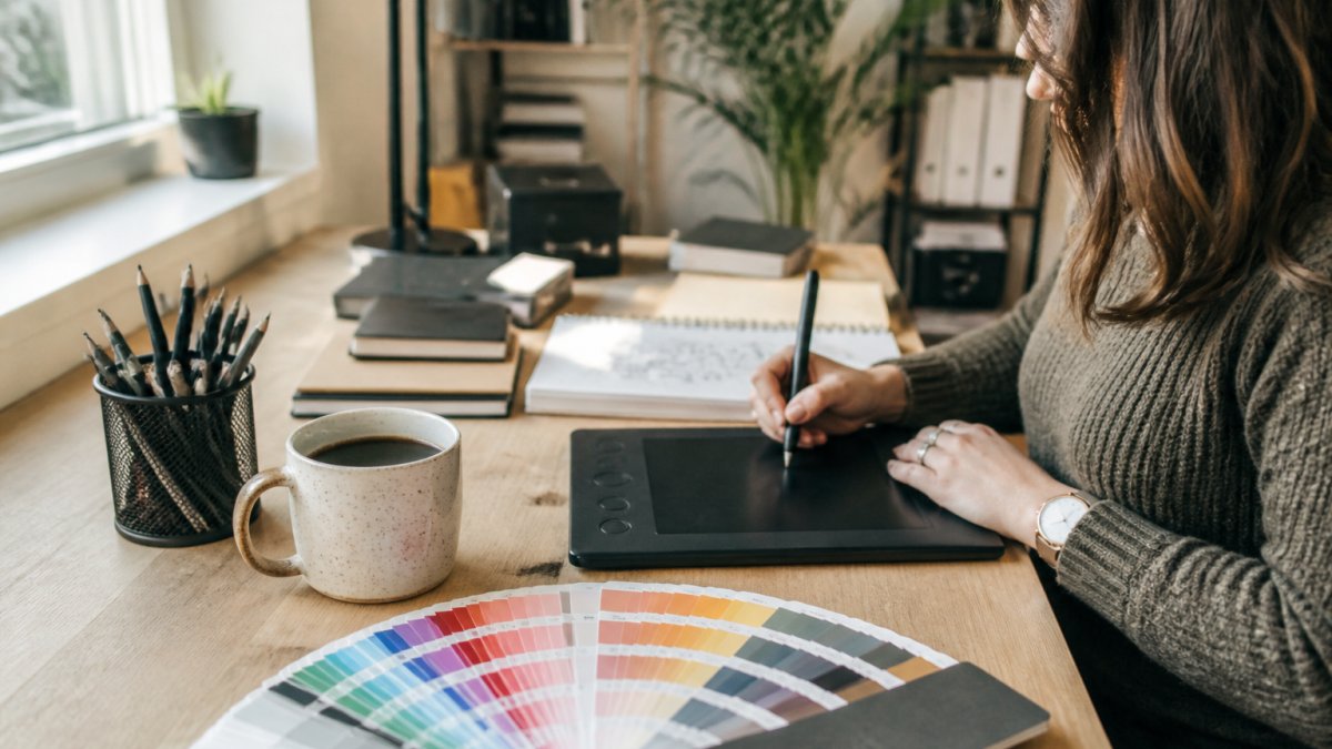 A graphic designer's desk with a drawing tablet, stylus, Pantone color guide, and a cup of coffee in a bright home studio