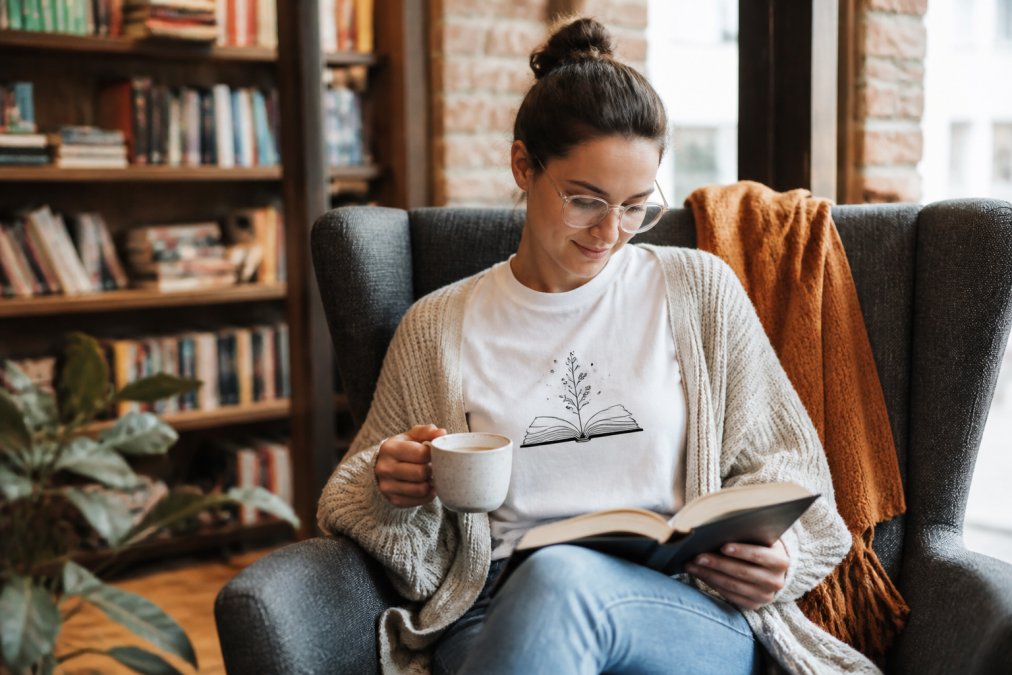 Person wearing a unique graphic shirt for bookworms while reading in a cozy nook with coffee and a cardigan