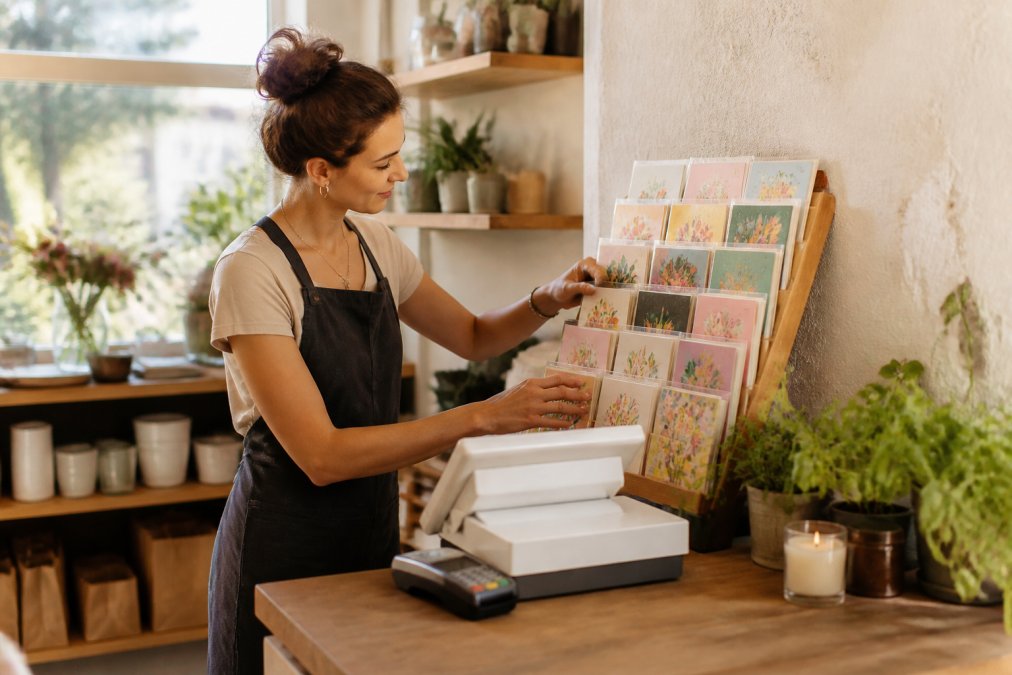 Boutique owner arranging unique greeting cards for boutique shops on a wooden display rack by the register
