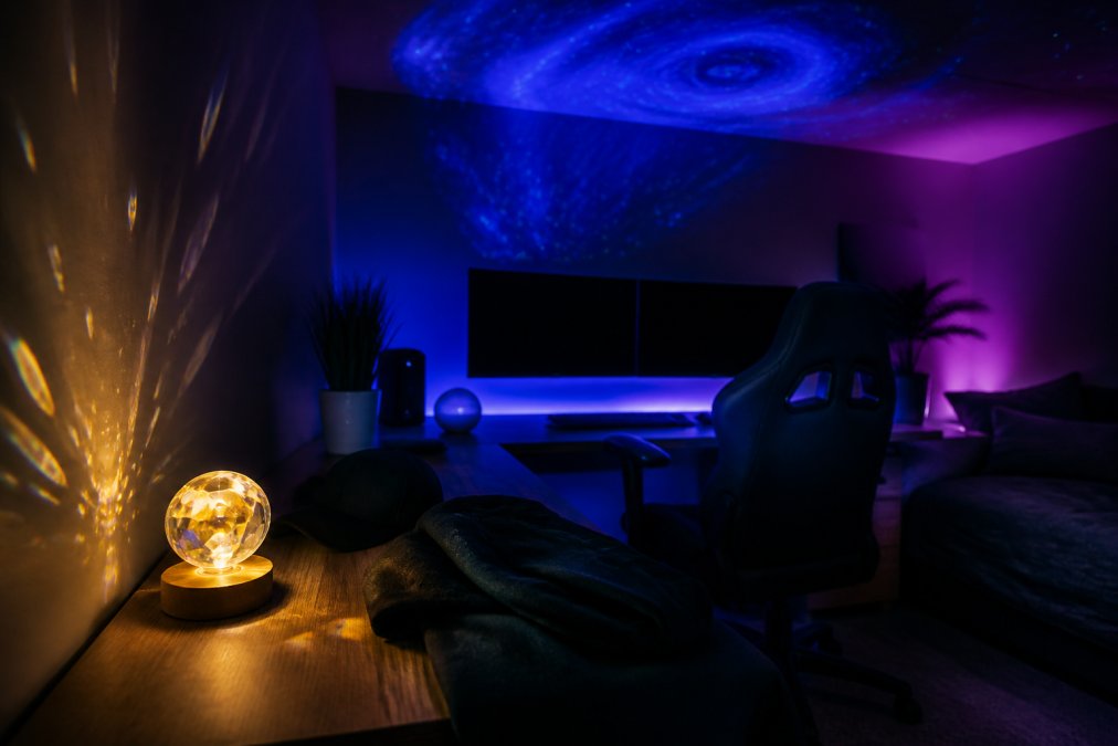 A dark gaming room glowing with deep blue and purple RGB backlighting behind dual monitors, a galaxy projector casting swirling star patterns across the ceiling, and a crystal sphere night light on the corner of a clean wooden desk casting warm prismatic reflections