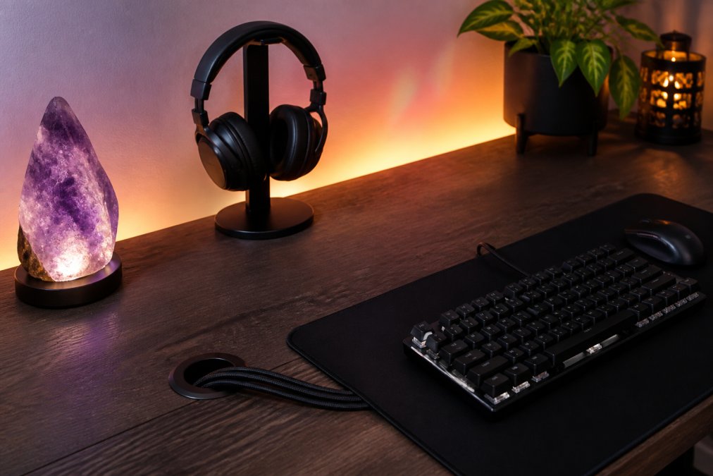 A clean gaming desk surface with a glowing crystal night light, a headset mounted on a side stand, neatly bundled cables routed through a desk grommet, and a minimalist keyboard and mouse on a wide desk mat under warm RGB backlighting