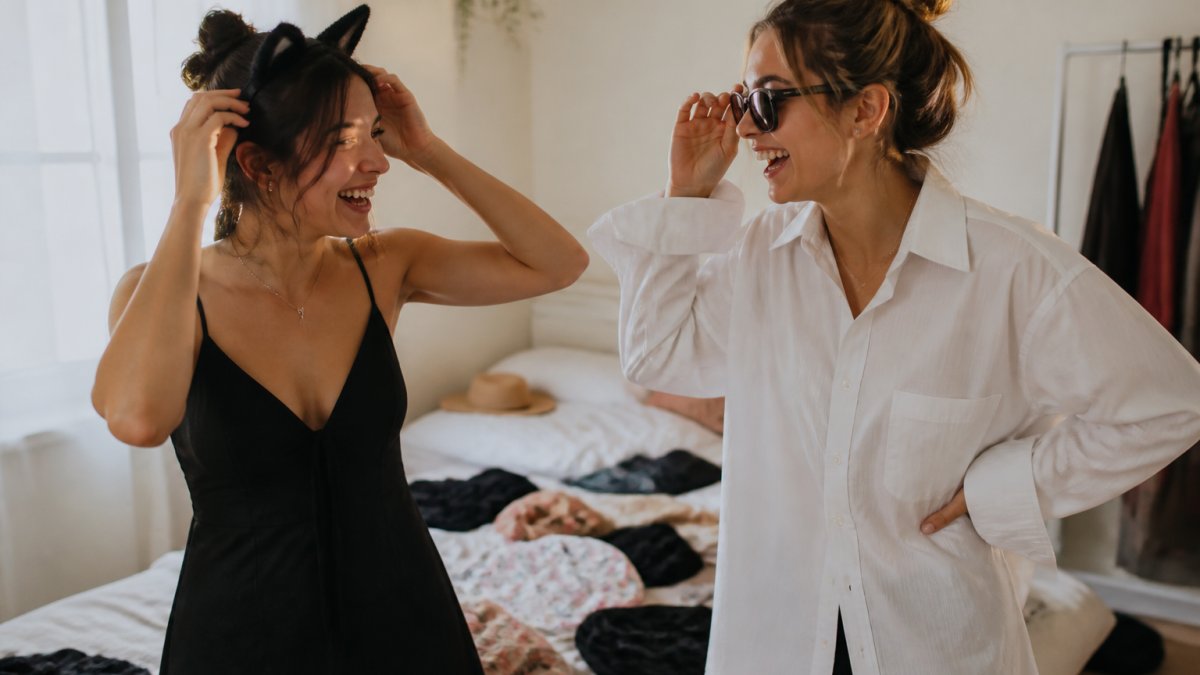 Two friends in their twenties laughing in a bedroom, one wearing a black dress with cat ears and the other in an oversized white button-down with sunglasses, clothes scattered on the bed behind them as they put together no-sew costumes