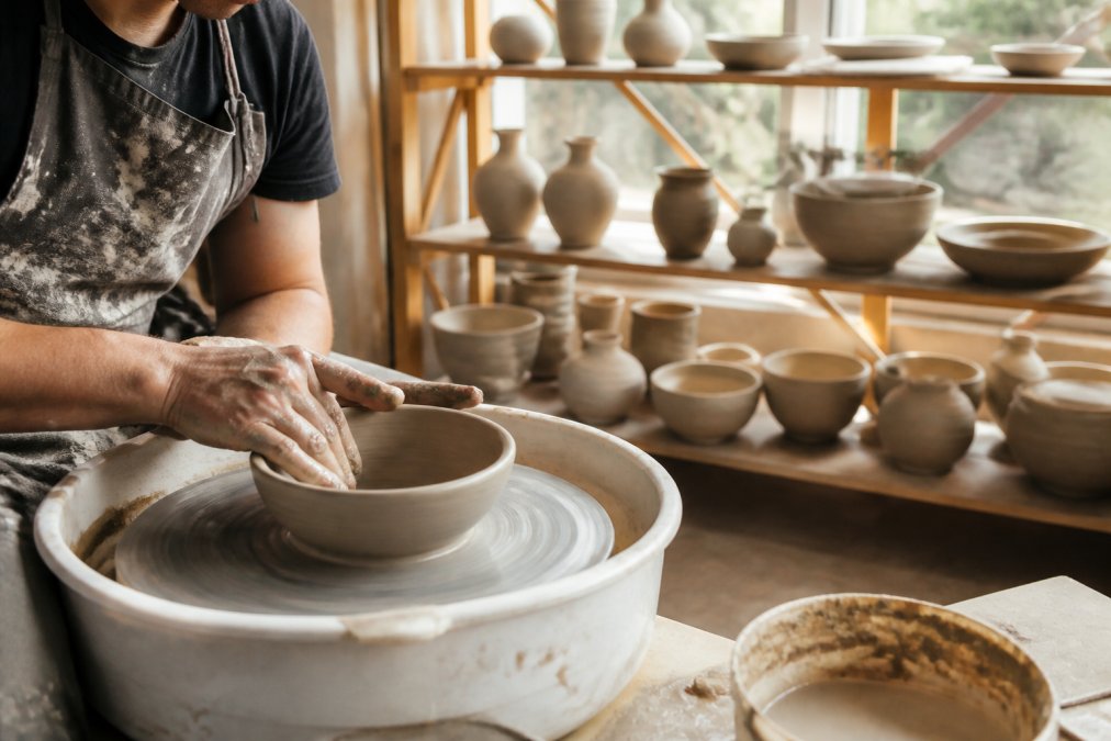 A close-up of a potter's hands shaping a clay bowl on a spinning wheel in a sunlit studio, with finished ceramic pieces resting on wooden shelves behind