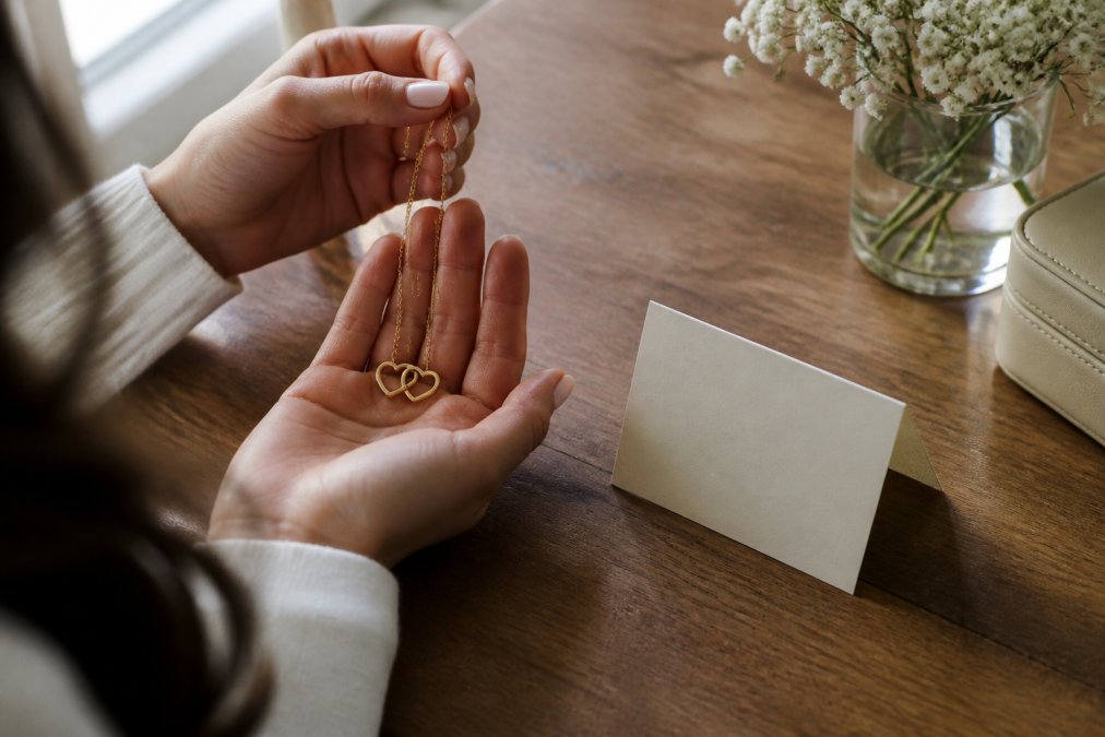 A woman's hands holding an elegant gold necklace with a small folded message card resting beside it on a wooden table, soft natural window light