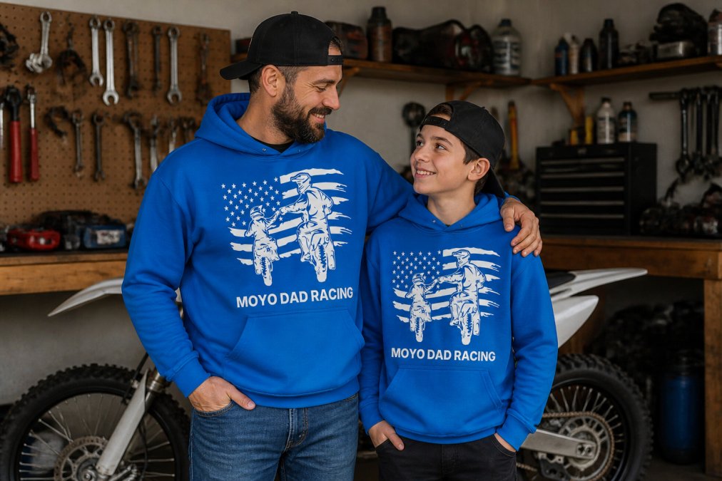 Father and teenage son wearing matching motocross-themed hoodies and tees standing in a garage workshop near a dirt bike, with tools and workbench visible in the background, natural overhead lighting