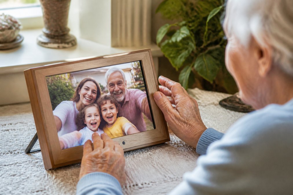 Close-up of an aging parent's hands gently touching a digital photo frame displaying vibrant family photos, with grandchildren's smiling faces visible on the bright screen, warm natural light streaming through a window