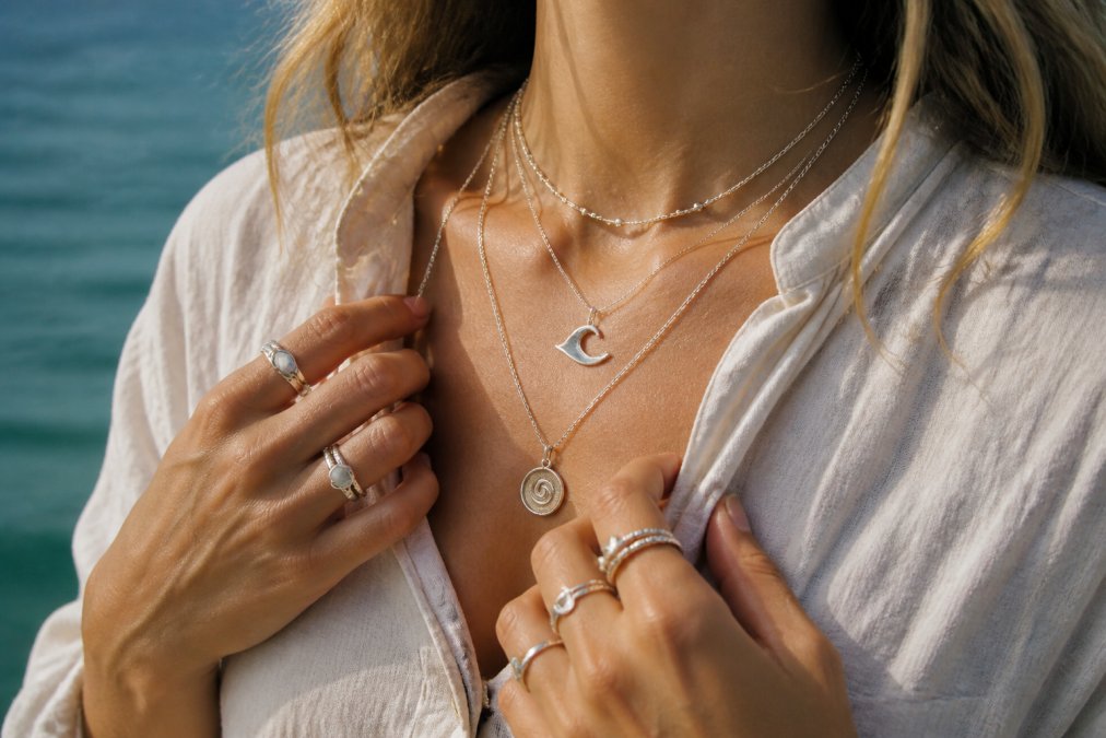 Close-up of a woman's hands and neck wearing layered handmade silver necklaces and a stack of silver rings, warm natural light, casual linen clothing, ocean-toned background