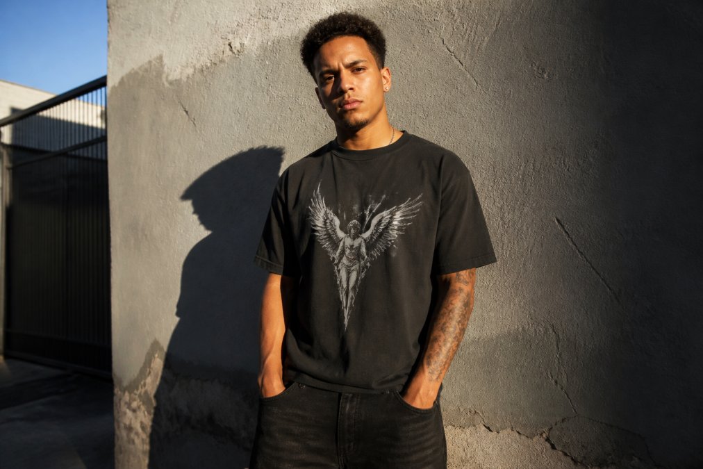 A young man in a well-fitted heavyweight graphic t-shirt and dark jeans standing against a textured urban wall, direct eye contact with the camera, late afternoon golden light casting long shadows