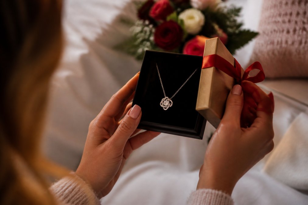 Close-up of a woman's hands opening a wrapped gift box containing a personalized engraved love knot necklace, with soft warm lighting and a romantic bedroom setting with flowers visible in the background
