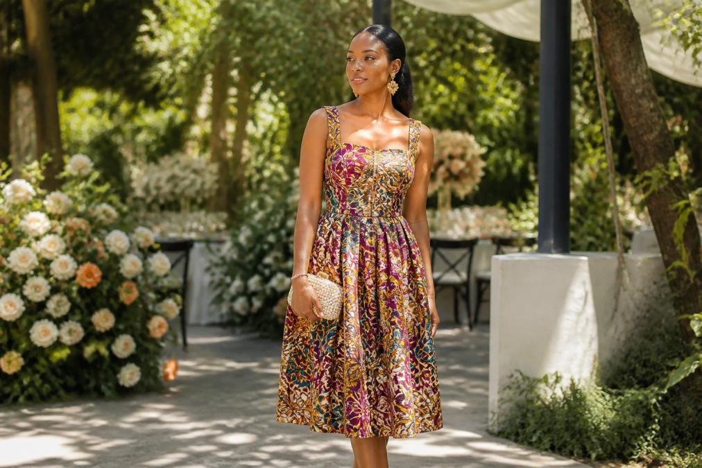 A woman in a vibrant artisan-printed midi dress with structured bodice, wearing gold statement earrings and holding a small beaded clutch, standing in dappled sunlight at an outdoor garden wedding venue surrounded by floral arrangements