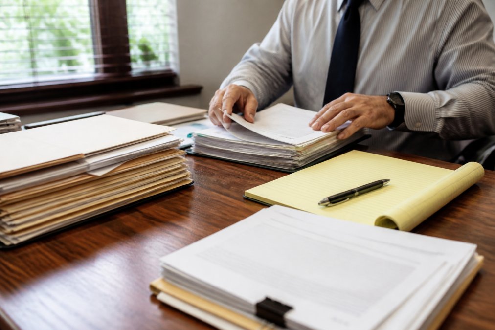 Professional gathering employment documents and emails on a desk, reviewing personnel records and correspondence with a notepad and pen nearby, natural office lighting