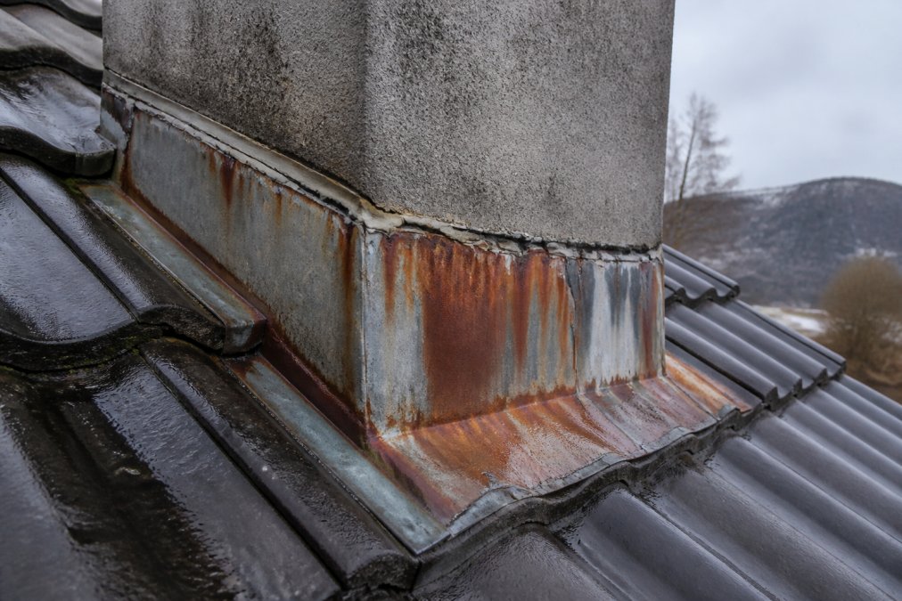 Close-up of a chimney base on a Norwegian pitched roof showing rust-streaked metal flashing, cracked sealant along the edges, and wet dark roof tiles immediately surrounding the chimney, photographed in overcast daylight