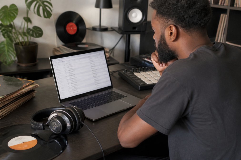 Independent music producer at desk with laptop displaying streaming analytics dashboard showing metadata fields and ISRC codes, headphones and vinyl records nearby in home studio
