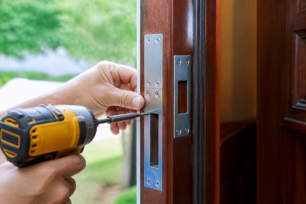 Close-up of hands installing a galvanized steel strike plate and reinforcement hardware on a wooden door frame, showing the drill, screws, and positioning of the components