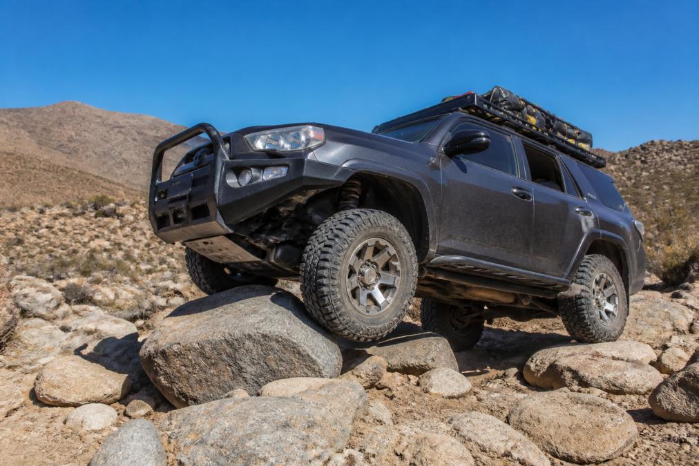SUV navigating rocky, uneven terrain in desert landscape with visible suspension articulation and wheel travel as vehicle climbs over large boulders