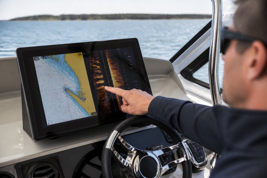 Modern boat helm equipped with a bright, high-resolution touchscreen chartplotter displaying detailed navigation data with side imaging sonar overlay, clear visibility in daylight with the captain's hand pointing at a specific waypoint