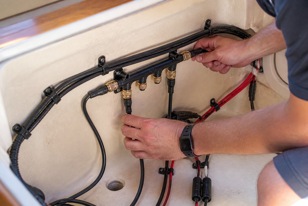 Hands carefully routing marine-grade wiring harness through boat cabin with proper strain relief clamps and protective conduit tubing, bright cabin lighting showing organized cable management
