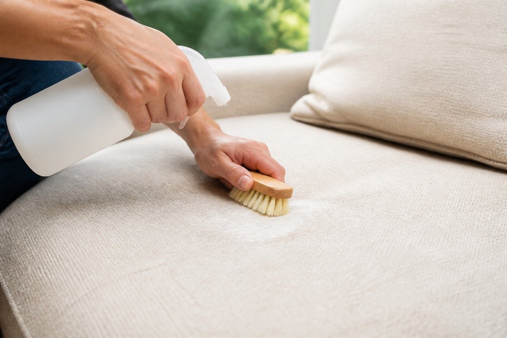 Close-up of hands applying cleaning solution to upholstery fabric with soft-bristled brush, demonstrating proper spot treatment technique on light-colored sofa with natural daylight