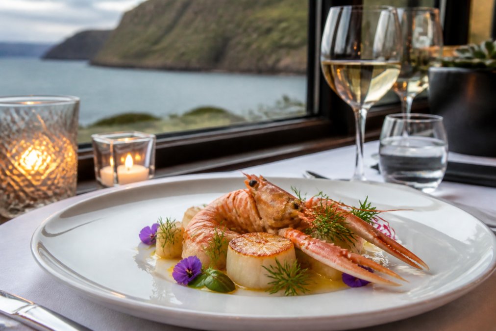 An elegantly plated seafood dish featuring fresh local langoustines and hand-dived scallops, garnished with delicate herbs and edible flowers, on fine white porcelain with soft candlelight reflecting off glassware and ocean views visible through the restaurant window