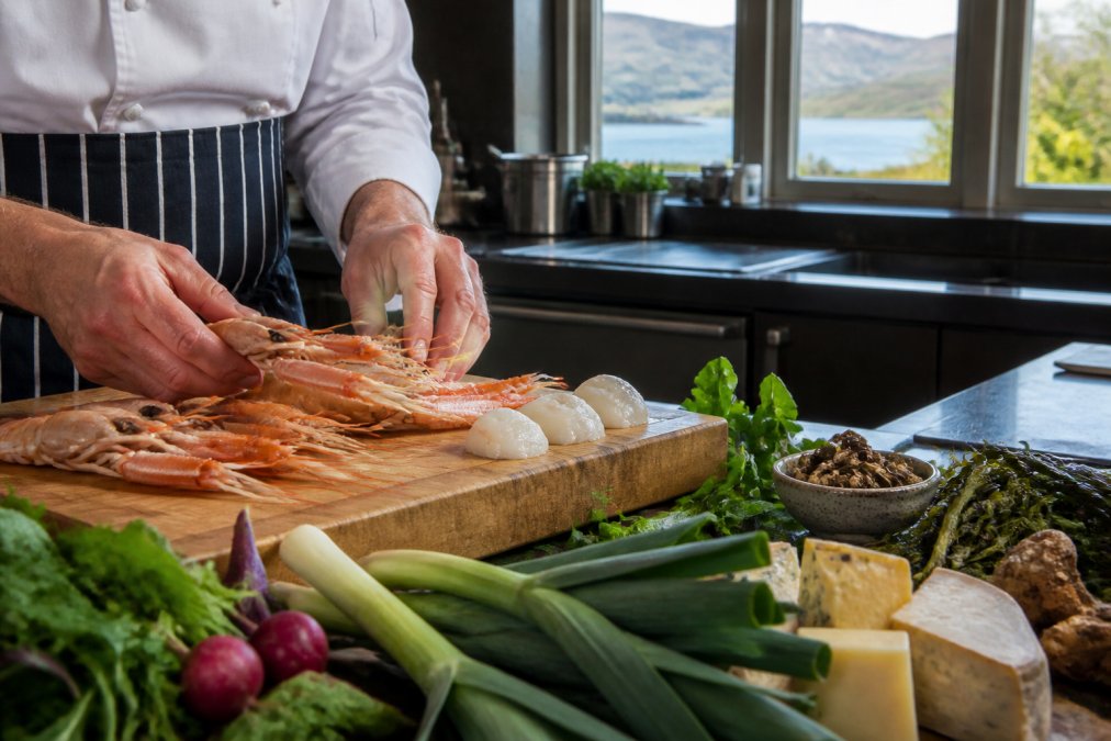 A chef's hands expertly preparing fresh local langoustines and hand-dived scallops on a wooden cutting board, with Highland vegetables, artisan cheeses, and foraged ingredients arranged nearby in a professional kitchen with natural light streaming through large windows