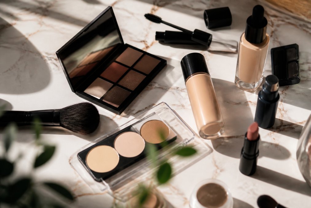 Close-up of diverse makeup products including foundation bottles, eyeshadow palettes, and mascara arranged on a light marble surface with natural sunlight streaming across the products